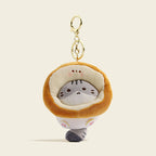 A plush gray cat bag charm inside a golden-brown Toast-shaped costume with embroidered whiskers and pink paw details. It is attached to a gold lobster clasp and keyring, designed as a cute and trendy accessory for bags or keychains.