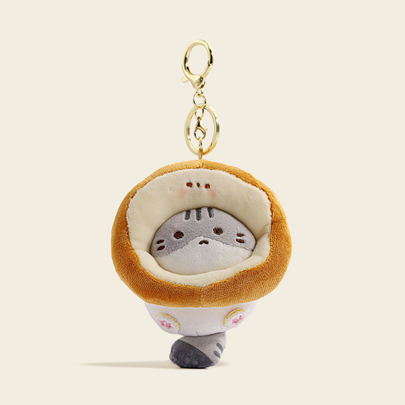 A plush gray cat bag charm inside a golden-brown Toast-shaped costume with embroidered whiskers and pink paw details. It is attached to a gold lobster clasp and keyring, designed as a cute and trendy accessory for bags or keychains.