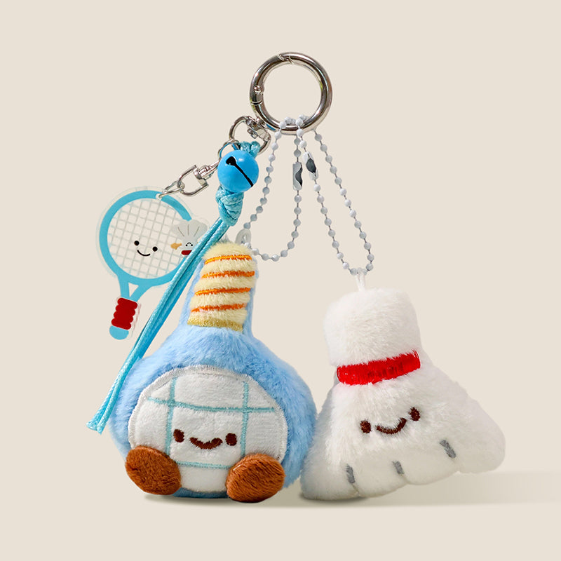 A plush keychain set with a blue badminton racket and a white shuttlecock, both with smiling faces, hanging from a keyring with a blue bell and a small acrylic racket charm.