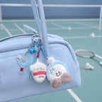 Two plush badminton shuttlecock keychains hanging from a light blue handbag; one wears a red headband, the other a blue hat with pom-poms, accompanied by a small acrylic badminton racket charm.