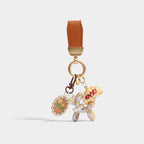 A keychain featuring a brown leather strap with gold-tone hardware, from which hang three charms: a brown scalloped tag with yellow flowers and the word 'flower', a pearlescent iridescent balloon dog, and a miniature croissant with red berry details.