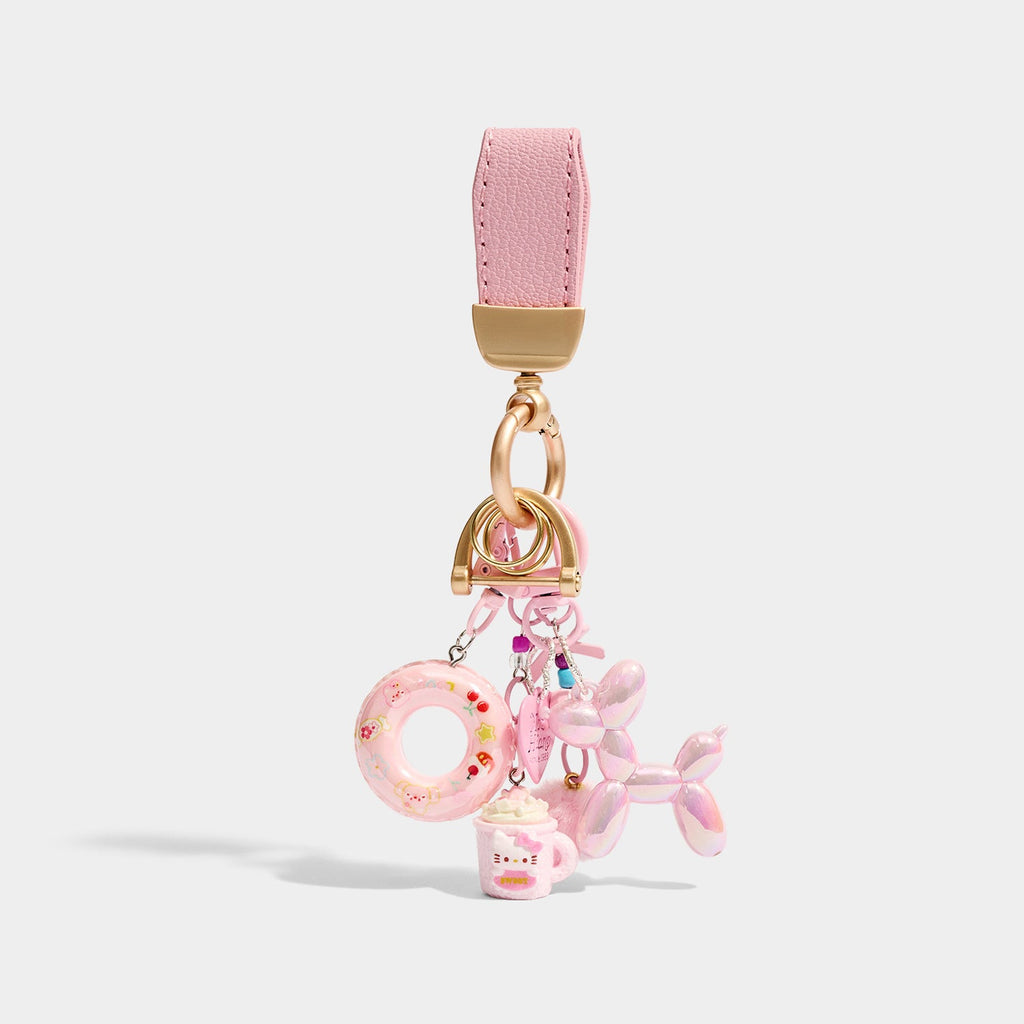 A keychain with a pink leather strap and gold-tone hardware, holding a set of charms including a pink inflatable ring with floral patterns, a pink Hello Kitty-themed mug, and a shiny pink iridescent balloon dog.