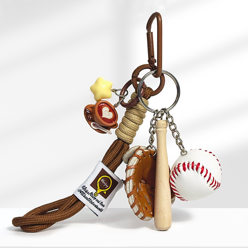 A baseball-themed keychain with a brown carabiner clip, featuring a yellow star charm, a brown coffee cup charm with a white heart design, a brown leather baseball glove, a wooden baseball bat, and a white baseball with red stitching, attached to a braided rope strap.