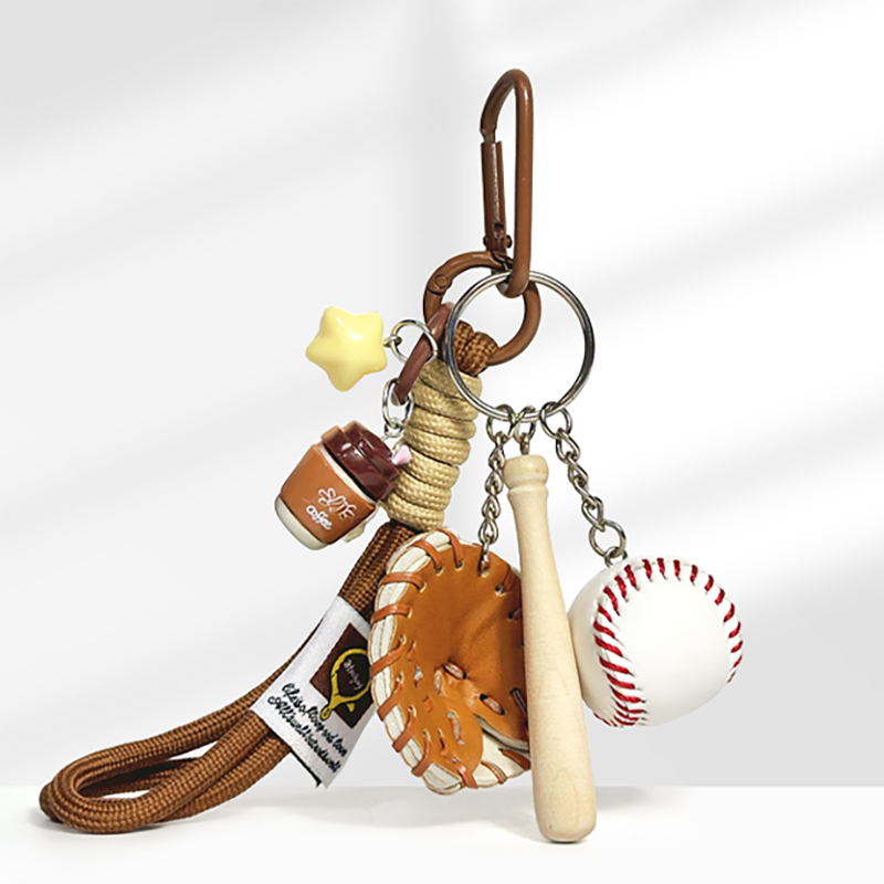 A baseball-themed keychain with a brown carabiner clip, featuring a yellow star charm, a brown lidded coffee cup charm with "cafe" text, a brown leather baseball glove, a wooden baseball bat, and a white baseball with red stitching, attached to a braided rope strap.