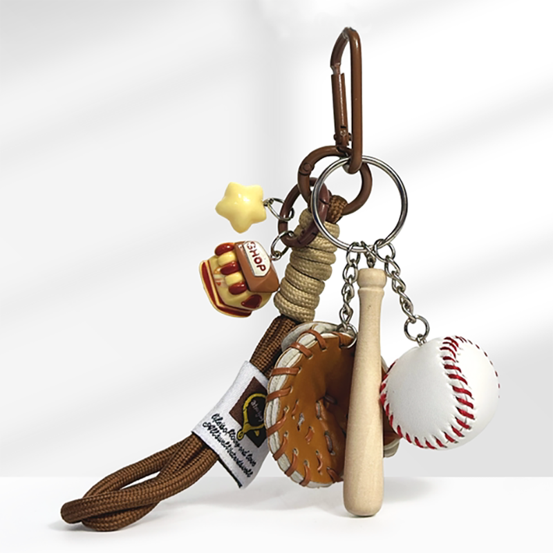 A baseball-themed keychain with a brown carabiner clip, featuring a yellow star charm, a brown hot dog charm with "HOTDOG" text, a brown leather baseball glove, a wooden baseball bat, and a white baseball with red stitching, attached to a braided rope strap.