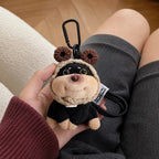 A plush dog bag charm with brown crocheted bear ears on its hood, held in a person's hand against a background of grey shorts. The charm features a black carabiner clip and a white tag with text, showcasing its detailed design as a trendy bag accessory.