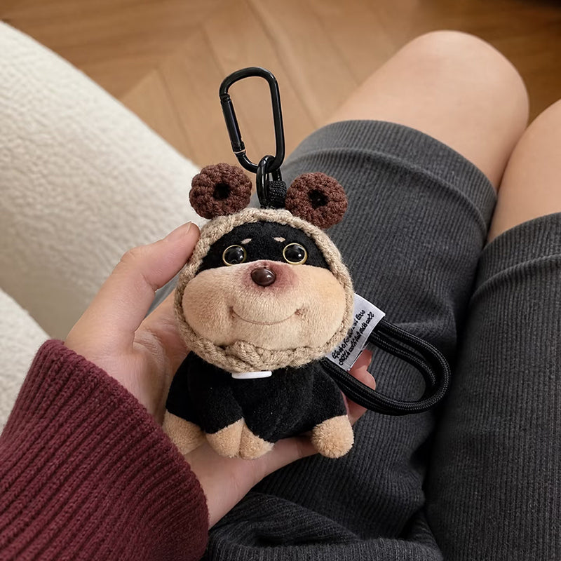 A plush dog bag charm with brown crocheted bear ears on its hood, held in a person's hand against a background of grey shorts. The charm features a black carabiner clip and a white tag with text, showcasing its detailed design as a trendy bag accessory.