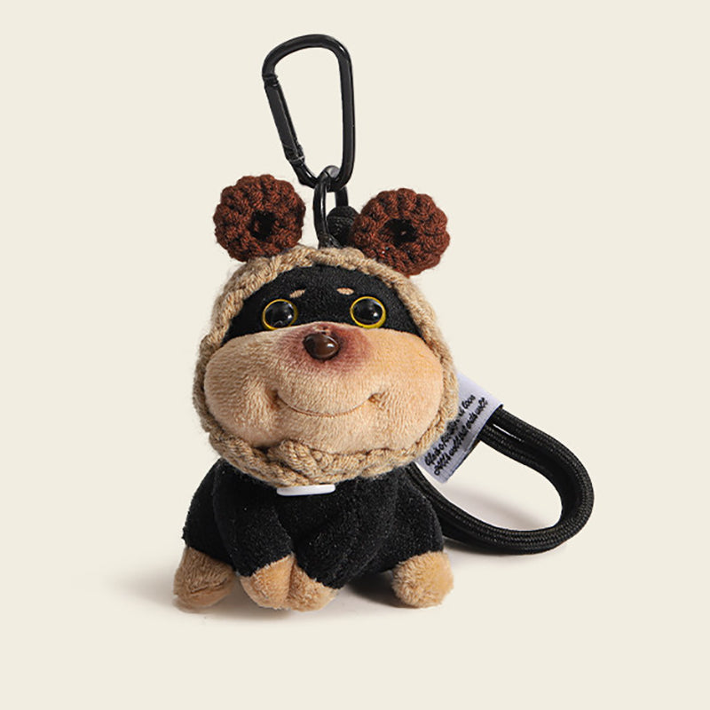 A plush dog bag charm with brown crocheted bear ears on its hood, attached to a black carabiner clip. The charm has a smiling face and is dressed in a black outfit, making it a charming and whimsical addition to any bag or keychain.