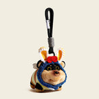 A plush bee-dog bag charm in a blue hood with white and yellow crocheted bunny ears and a red flower, attached to a black carabiner clip with a white tag. The charm has a smiling face and a white button on its chest, making it a charming and whimsical addition to any bag or keychain.