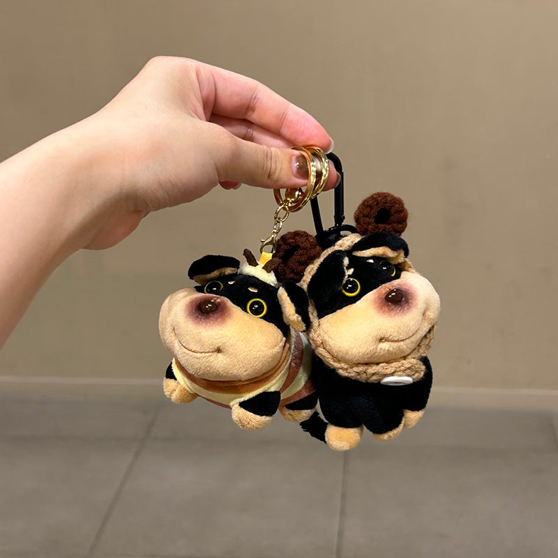 Two plush bee-dog bag charms held in a person's hand, one in a bee costume and the other in a black outfit with brown crocheted antennae. Both charms feature smiling faces and are attached to carabiner clips, showcasing their detailed design as trendy bag accessories.