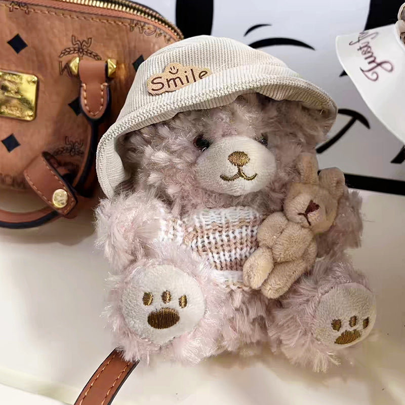 A close-up of a light brown teddy bear plush keychain wearing a beige corduroy hat with a "Smile" patch and a beige and white striped knit sweater, holding a small beige rabbit plush, with a brown leather bag in the background.