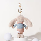 A beige plush rabbit keychain with long floppy ears, wearing a blue polka-dot shirt and a pink corduroy vest, shown from the back, attached to a gold-tone metal carabiner and keyring, displayed on a white pedestal.