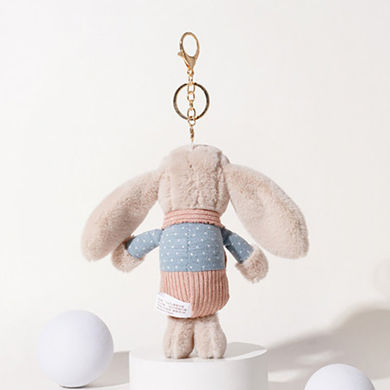 A beige plush rabbit keychain with long floppy ears, wearing a blue polka-dot shirt and a pink corduroy vest, shown from the back, attached to a gold-tone metal carabiner and keyring, displayed on a white pedestal.