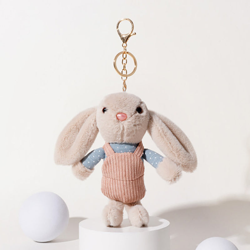 A beige plush rabbit keychain with long floppy ears, black embroidered eyes, and a pink nose, wearing a blue polka-dot shirt and a pink corduroy vest, attached to a gold-tone metal carabiner and keyring, displayed on a white pedestal.