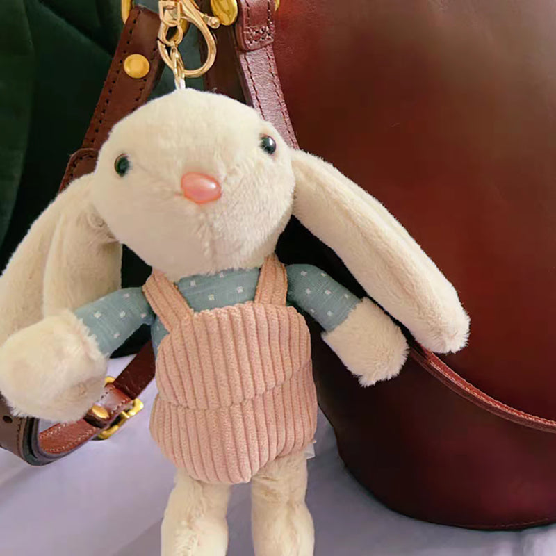 A beige plush rabbit keychain with long floppy ears, wearing a blue polka-dot shirt and a pink corduroy vest, attached to a gold-tone metal carabiner and hanging from a brown leather bag.