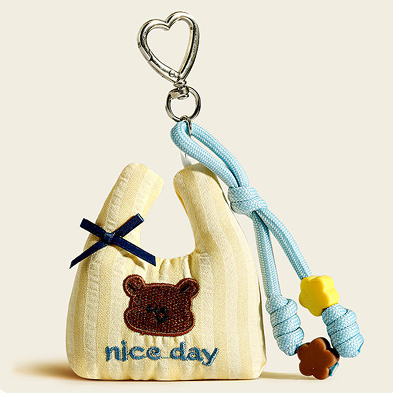 A beige striped fabric bag charm shaped like a mini tote, with an embroidered brown bear and "nice day" text, decorated with a blue bow and yellow/brown flower beads.