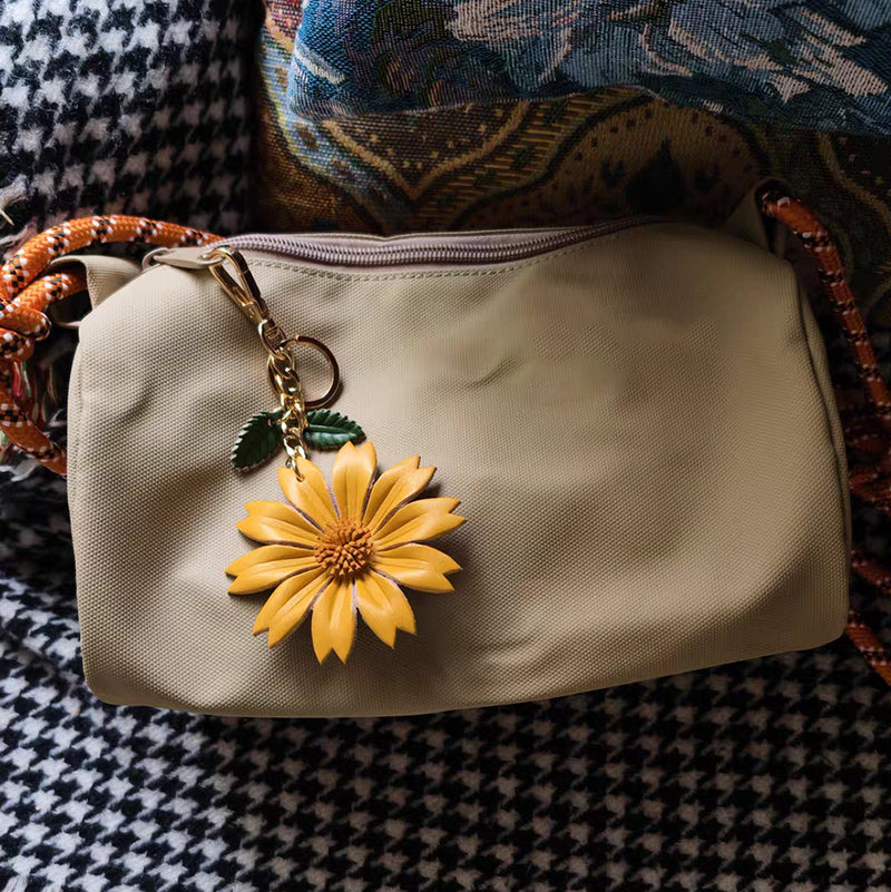 A beige fabric pouch adorned with a bright yellow leather daisy charm clipped onto its zipper pull. The accessory features green leaf accents and rests atop a patterned textile surface with houndstooth and floral motifs.