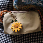 A beige fabric pouch adorned with a bright yellow leather daisy charm clipped onto its zipper pull. The accessory features green leaf accents and rests atop a patterned textile surface with houndstooth and floral motifs.