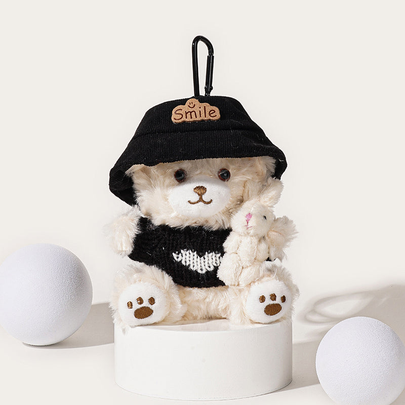 A front view of a white teddy bear plush keychain wearing a black bucket hat with a "Smile" patch and a black knit sweater with a white heart, holding a small white rabbit plush, attached to a black carabiner clip.