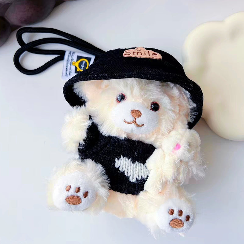 A white teddy bear plush keychain wearing a black bucket hat with a "Smile" patch and a black knit sweater with a white heart, holding a small white rabbit plush, with a black strap attached.