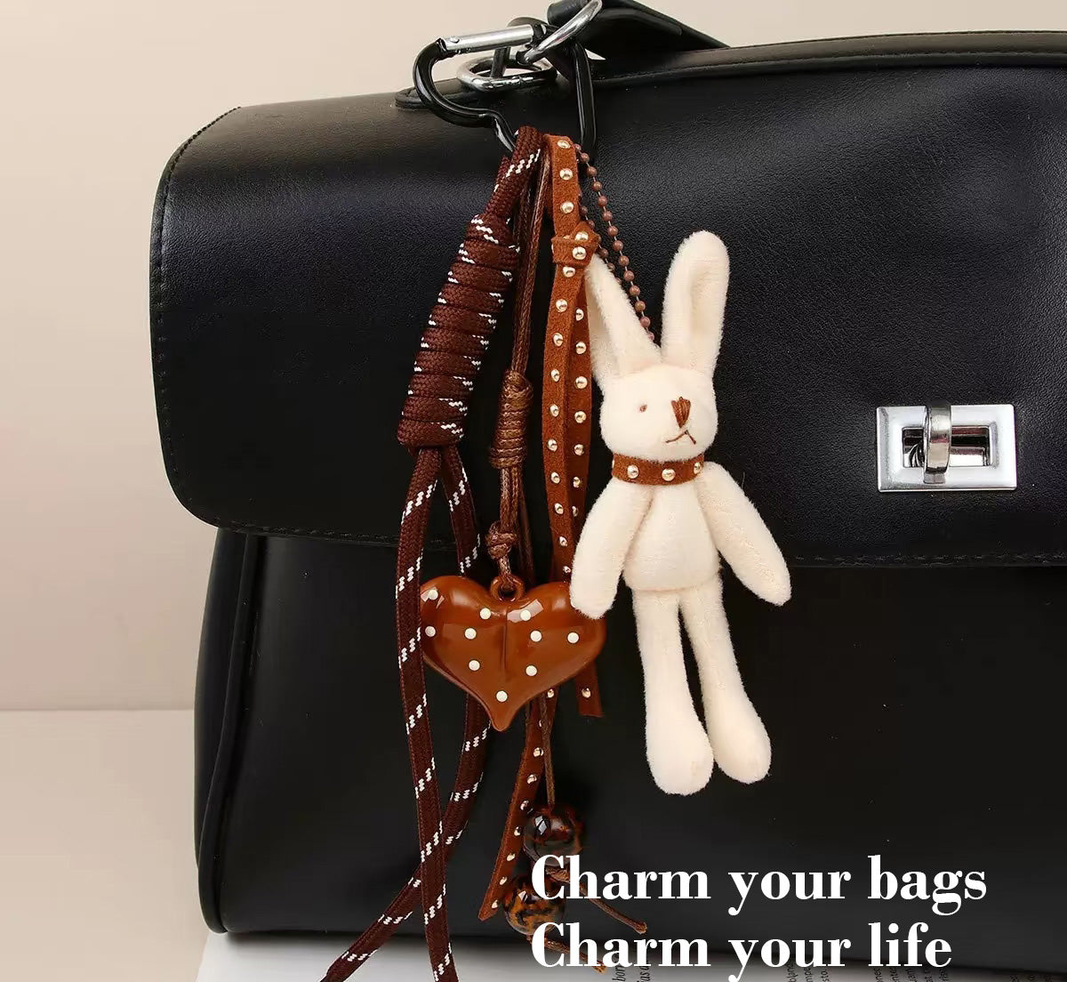 A black leather handbag with a silver-tone clasp, adorned with multiple charms: a white plush rabbit with brown collar, a brown heart-shaped pendant with white polka dots, and braided brown straps with metallic studs. The text overlay reads “Charm your bags, Charm your life.”