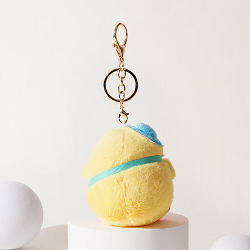 Back view of a yellow plush chick keychain with a blue hat and light blue ribbon, attached to a gold-tone metal carabiner and keyring, displayed on a white pedestal.
