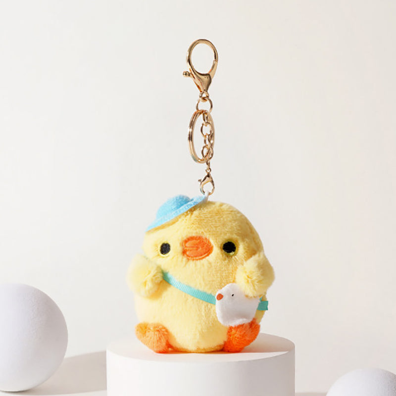 Front view of a yellow plush chick keychain wearing a blue hat and light blue ribbon, holding a small white bird charm, with black eyes and an orange beak, attached to a gold-tone metal carabiner and keyring.