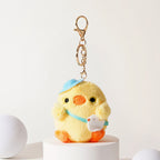 Front view of a yellow plush chick keychain wearing a blue hat and light blue ribbon, holding a small white bird charm, with black eyes and an orange beak, attached to a gold-tone metal carabiner and keyring.