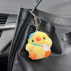 A yellow plush chick keychain wearing a blue hat and a light blue ribbon, holding a small white bird charm, attached to a gold-tone metal keyring and hanging from a black leather bag in a car interior.