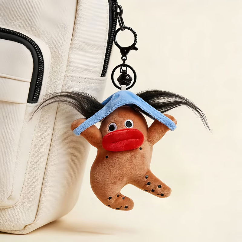 A plush gingerbread man bag charm with a blue hat, black pigtails, and a red mouth, attached to a white backpack with black zippers. The charm features a playful design with black dots on its body, making it a whimsical and trendy accessory for bags.