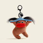 A plush gingerbread man bag charm with a blue hat, black pigtails, and a red mouth, attached to a black carabiner clip with a small bell. The charm has black dots on its body, designed as a cute and trendy accessory for bags or keychains.