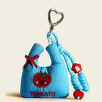 A front view of a light blue fabric bag charm shaped like a mini tote, with a red embroidered tomato and "TOMATO" text, accented with a red ribbon and red flower beads.