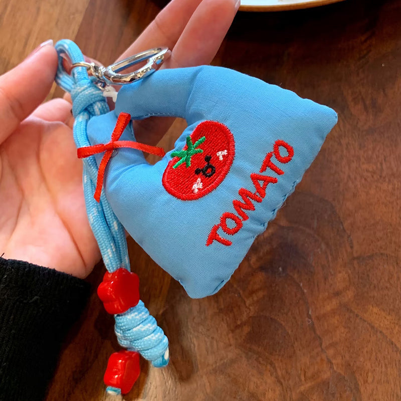 A hand holding a light blue fabric bag charm with a red embroidered tomato and the word "TOMATO", accented with a red ribbon and red flower beads on the strap.
