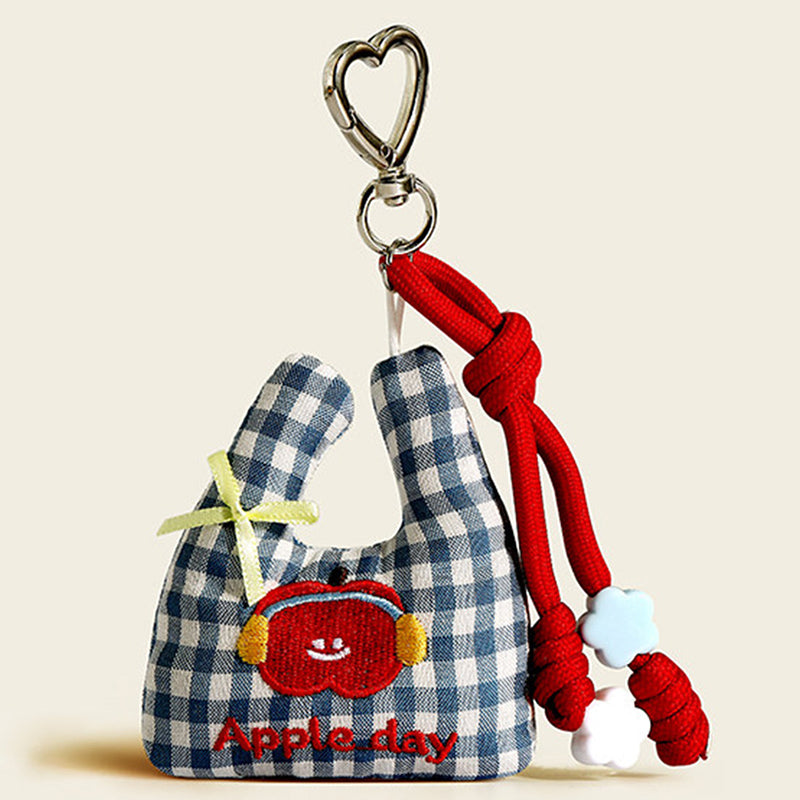 A blue and white plaid fabric bag charm shaped like a mini tote, featuring an embroidered red apple with earmuffs and "Apple day" text, accented with a yellow bow and white flower beads.