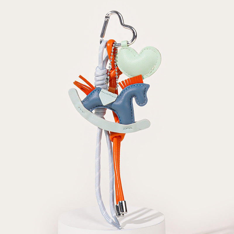 A pastel-colored leather keychain featuring a blue rocking horse and a mint green heart charm. It is decorated with orange tassels and hangs from grey and orange cords attached to a silver hook.