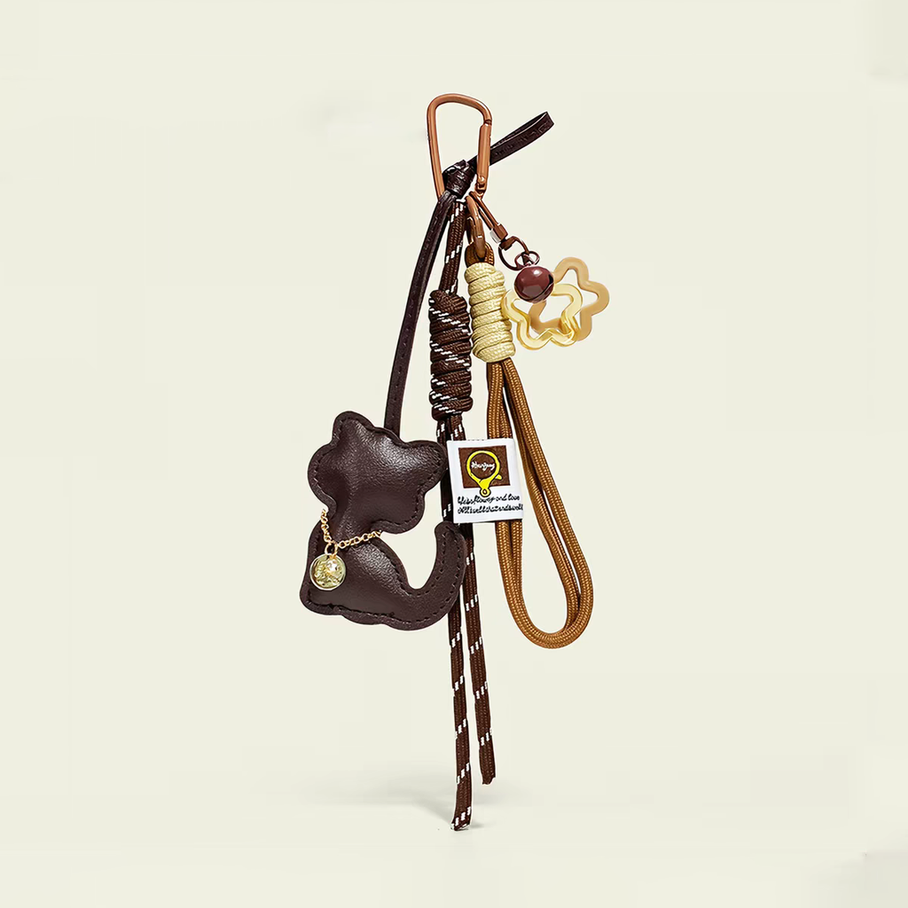 A brown leather cat-shaped charm with a small gold chain detail, attached to a copper-colored carabiner with woven cords and a star-shaped acrylic piece. A white tag with a logo is also attached.