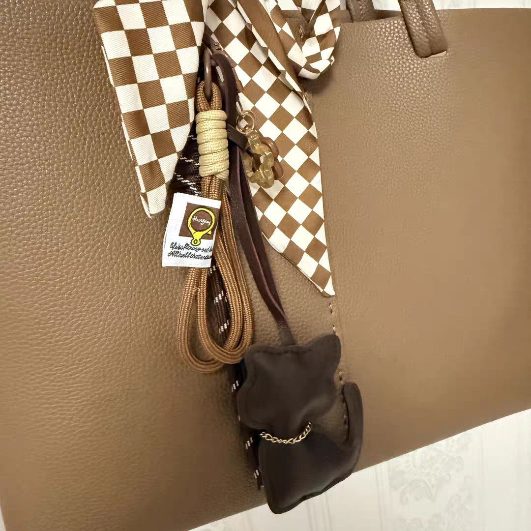 A brown leather cat-shaped charm hanging from a brown tote bag, styled with a brown-and-white checkered scarf. The charm is attached with woven cords and a copper carabiner, featuring a small gold chain and a white logo tag.