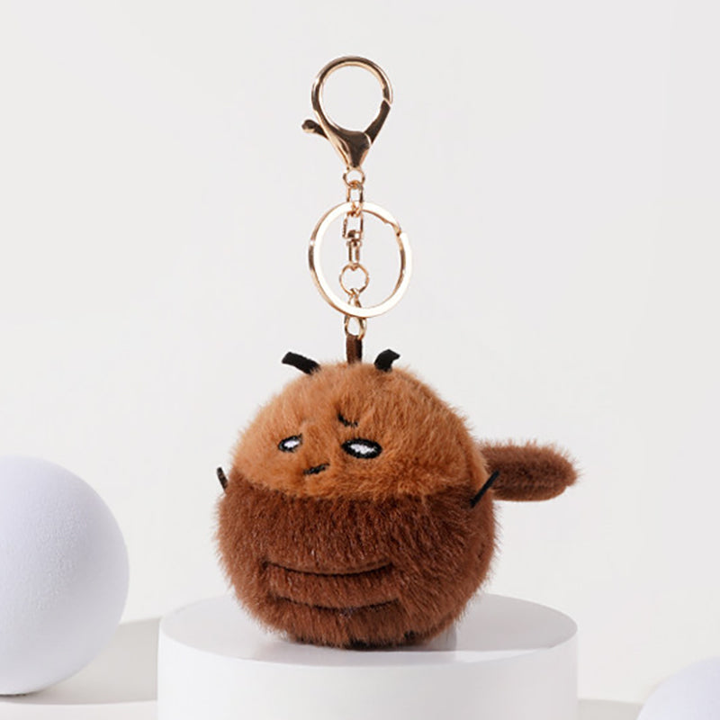 A side view of a brown plush cockroach keychain with embroidered details, black antennae, and a fuzzy body, attached to a gold-tone metal carabiner and keyring.