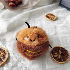 A brown plush cockroach toy with embroidered smiling face, pink cheeks, and black antennae, placed on a white textured fabric with dried orange slices nearby.