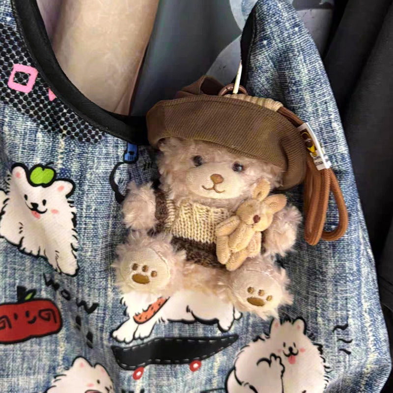 A light brown teddy bear plush keychain wearing a brown corduroy hat and a brown and beige striped knit sweater, holding a small beige rabbit plush, hanging from the strap of a blue denim bag with cartoon dog prints.