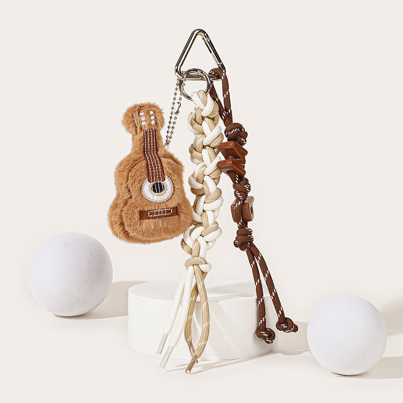 A brown plush guitar-shaped keychain with embroidered strings and soundhole, attached to a silver carabiner with a white and beige braided rope strap and brown decorative beads.