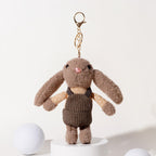 A brown plush rabbit keychain with long floppy ears, black embroidered eyes, and a pink nose, wearing a beige polka-dot shirt and a brown corduroy vest, attached to a gold-tone metal carabiner and keyring, displayed on a white pedestal.
