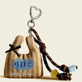 A brown striped fabric bag charm shaped like a mini tote, featuring an embroidered blue bread loaf and "BREAD DAY" text, accented with a black bow and yellow/brown flower beads.