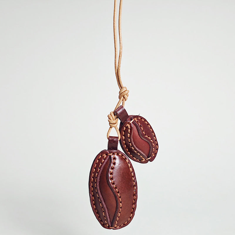 A close-up product shot of a double coffee bean keychain made from rich brown leather. It features detailed edge stitching and hangs vertically from a knotted leather strap.