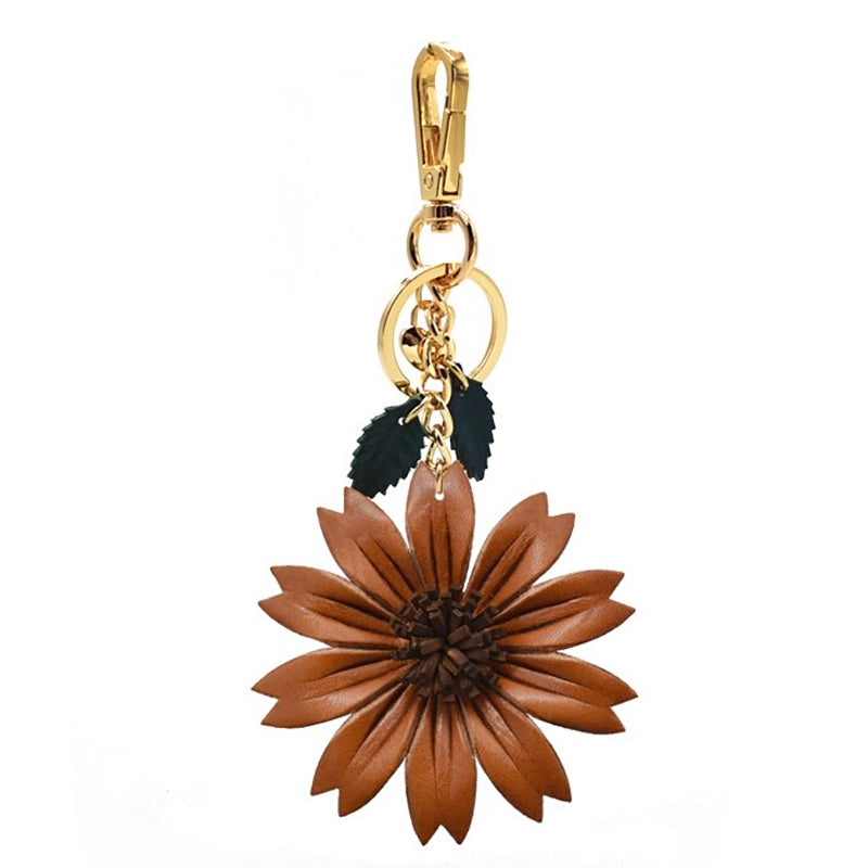 A close-up of a brown leather flower keychain suspended from a gold-tone lobster clasp. The flower has layered petals and is accompanied by small green leaf accents on a short chain.