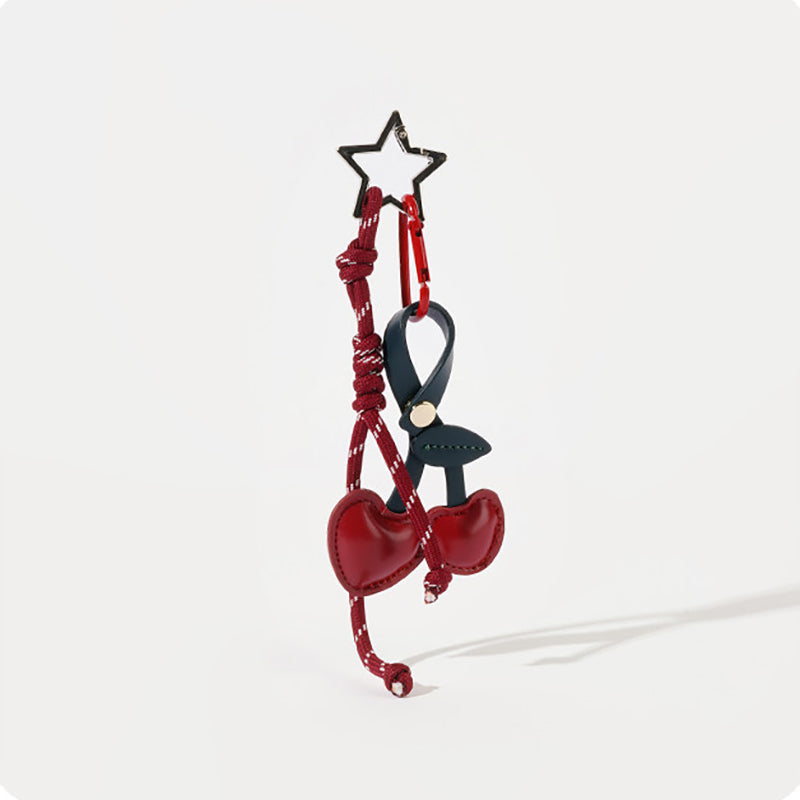 A burgundy leather cherry-shaped bag charm with a dark blue stem, attached to a matching burgundy braided rope strap. A black star-shaped clip is at the top, designed as a stylish and adorable accessory for bags or keychains.