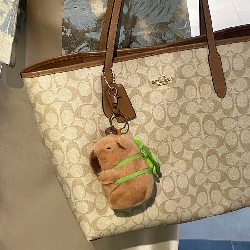 A plush capybara bag charm with a green backpack, attached to a brown leather Coach tote bag. The charm features a smiling face and is designed as a cute and trendy accessory for bags or backpacks.