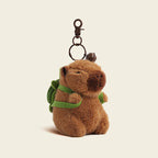 A plush capybara bag charm with a green backpack, attached to a brown carabiner clip. The charm features a smiling face and is designed as a cute and trendy accessory for bags or backpacks.