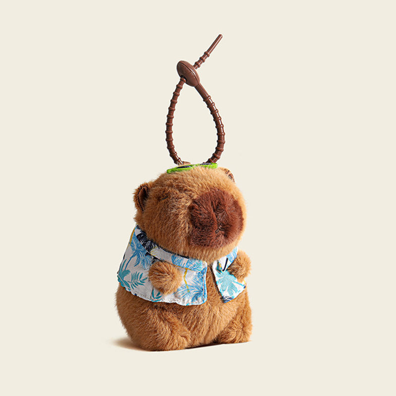 A plush capybara bag charm wearing a blue and white Hawaiian shirt with leaf patterns, attached to a brown carabiner clip. The charm has a smiling face and is designed as a cute and trendy accessory for bags or backpacks.