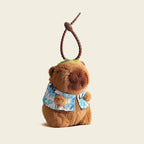 A plush capybara bag charm wearing a blue and white Hawaiian shirt with leaf patterns, attached to a brown carabiner clip. The charm has a smiling face and is designed as a cute and trendy accessory for bags or backpacks.